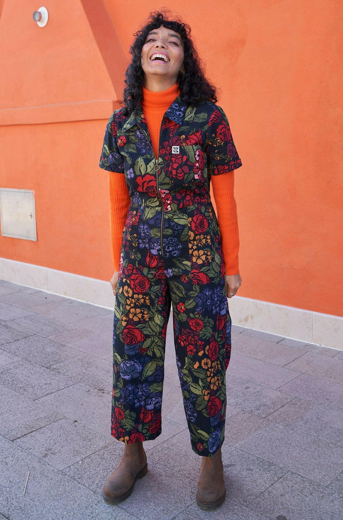A person wearing a multi-coloured floral jumpsuit with short sleeves and wide cropped legs, paired with an orange top and gray boots, standing in front of an orange wall.